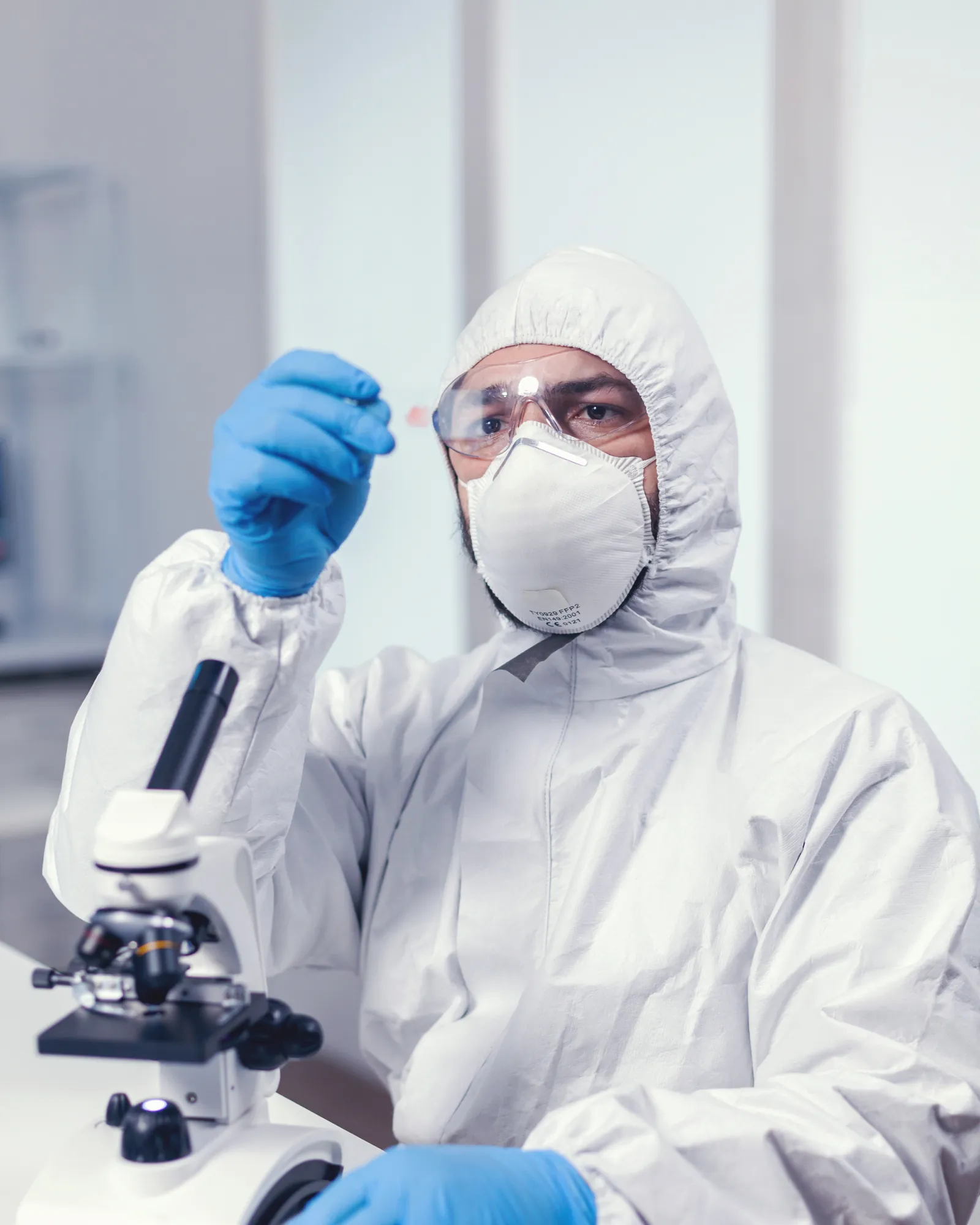 A scientist in a protective suit and respirator examining a sample beside a microscope